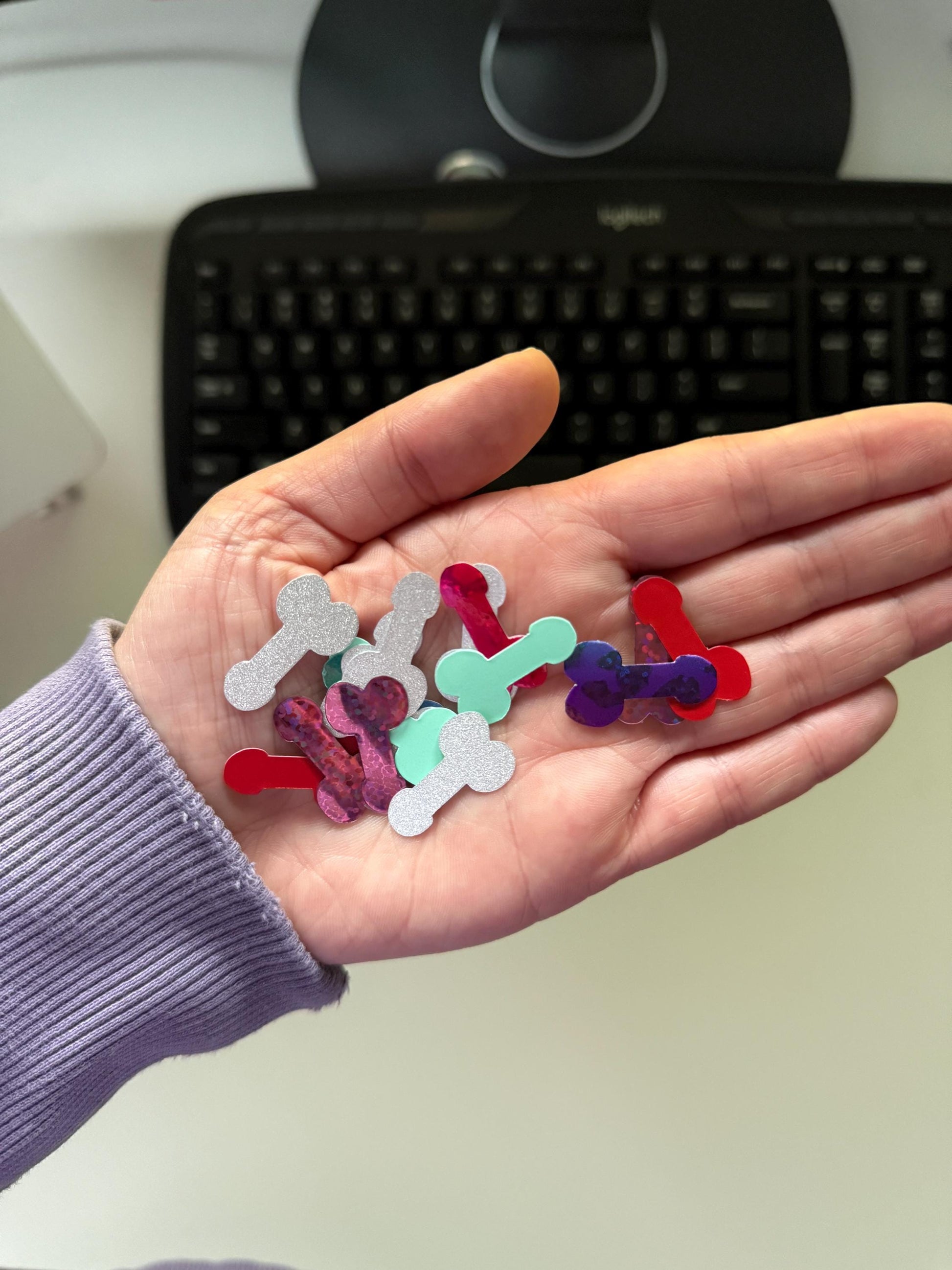 A collection of small, phallic-shaped sticker pieces in various finishes and colors, including silver glitter, solid red, mint green, and a dark purple with a subtle pattern, resting in a palm.