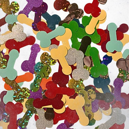A collection of scattered confetti pieces shaped like bones in various colors and finishes, including solid red, green, yellow, and light blue, alongside glittery purple, gold, silver, and multi-colored textured surfaces.